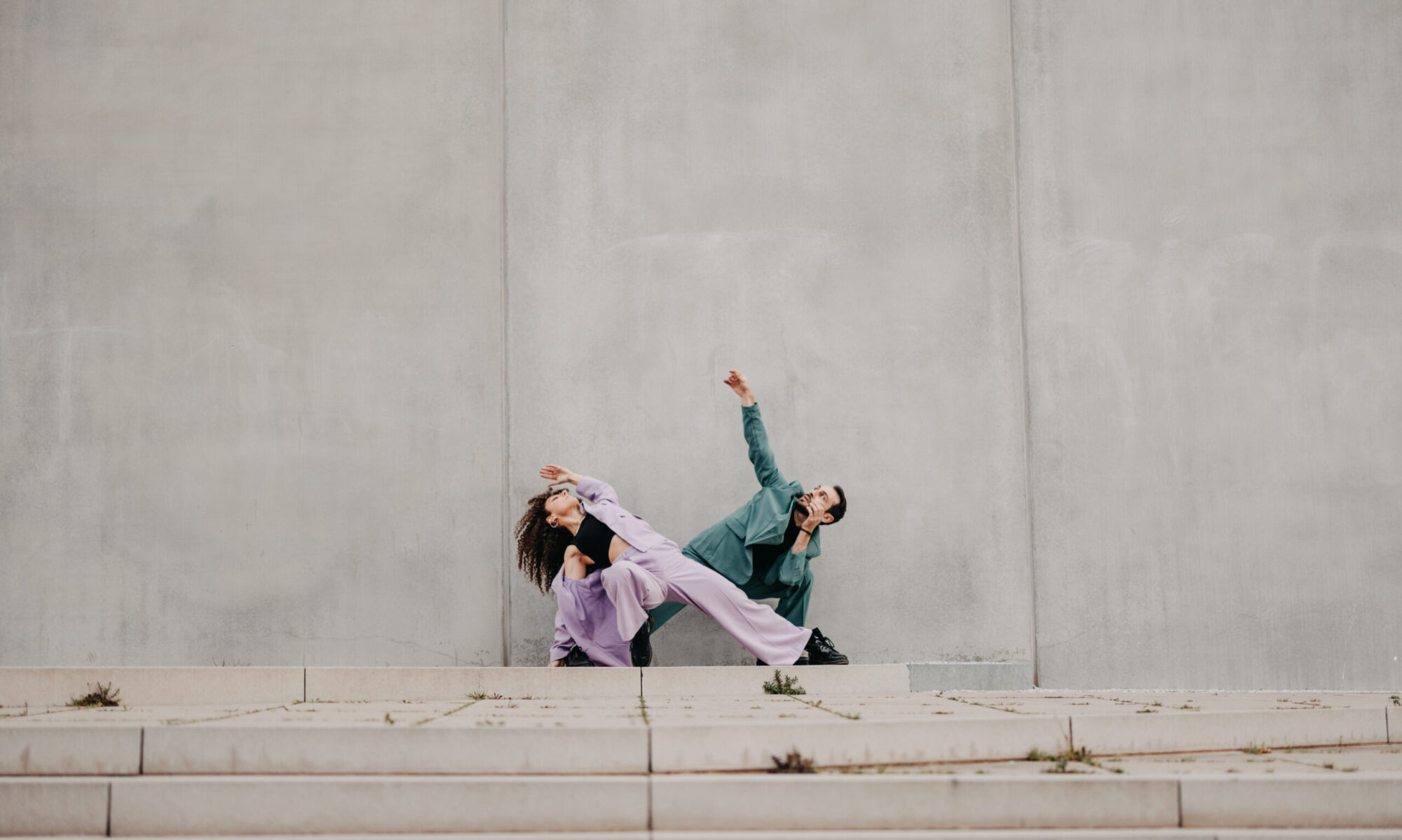 Un danseur et une danseuse en train de danser devant un mur en béton. Tous les deux portent des costumes